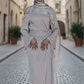 Woman in a long, light-colored dress with a headscarf standing on a cobblestone street.