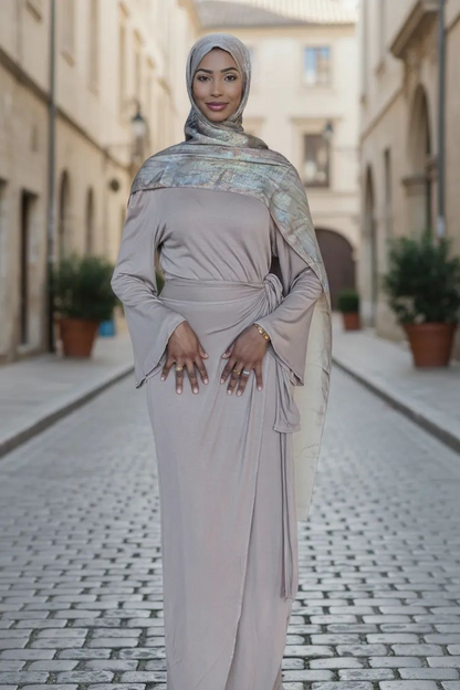 Woman in a long, light-colored dress with a headscarf standing on a cobblestone street.