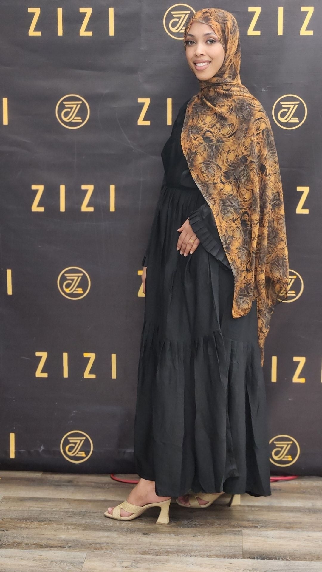 Woman in black dress and patterned hijab standing in front of a ZIZI branded backdrop