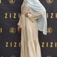 Woman in a beige dress with a light blue hijab standing in front of a ZIZI branded backdrop.