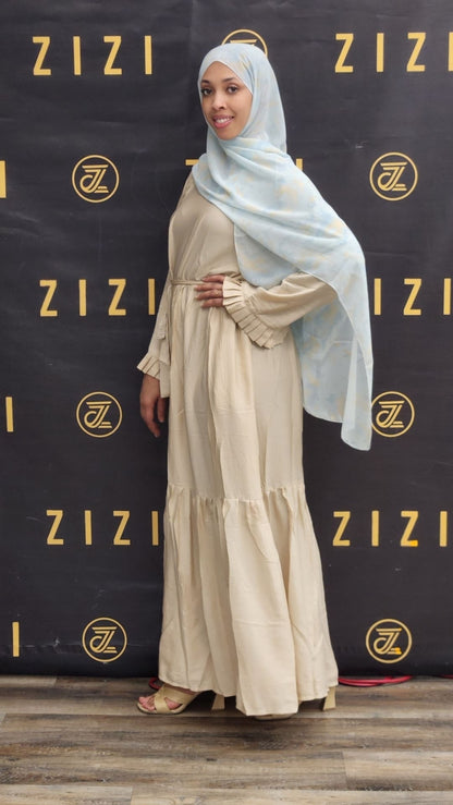 Woman in a beige dress with a light blue hijab standing in front of a ZIZI branded backdrop.