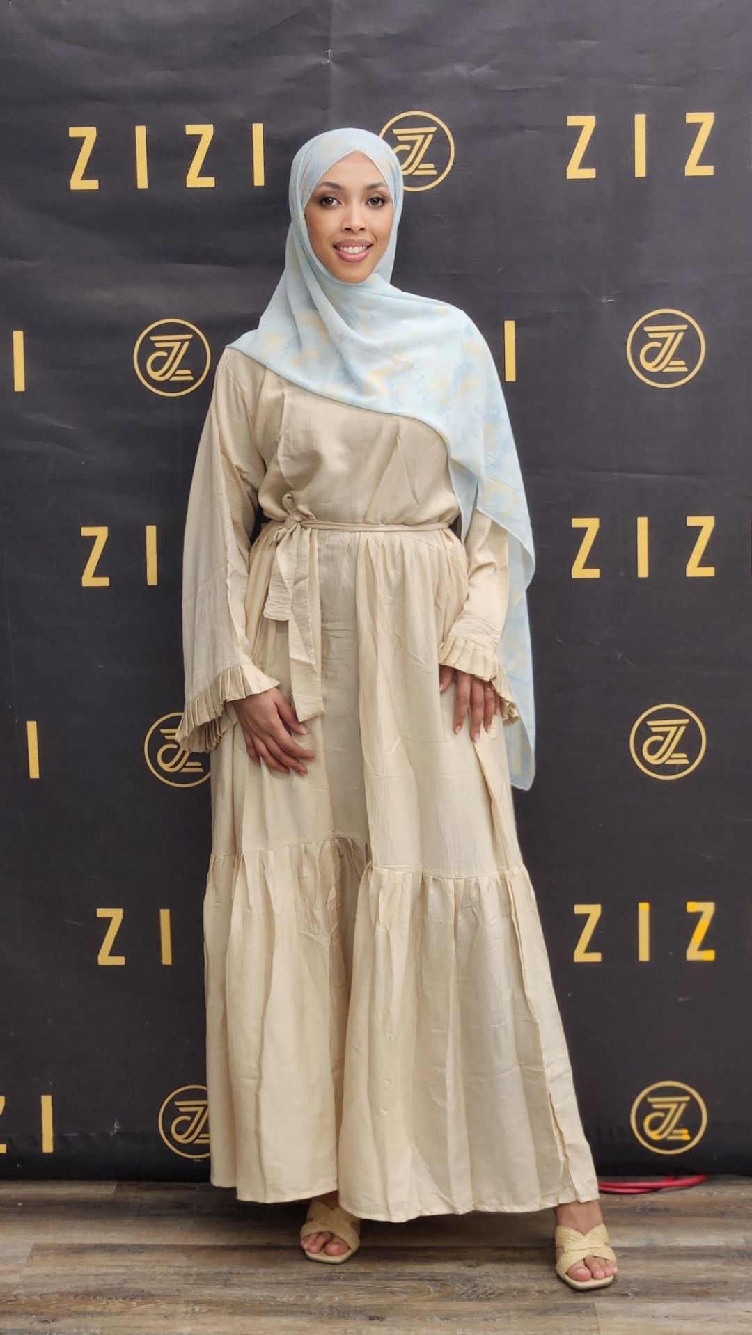 Woman in a beige dress with a light blue hijab standing in front of a branded backdrop.