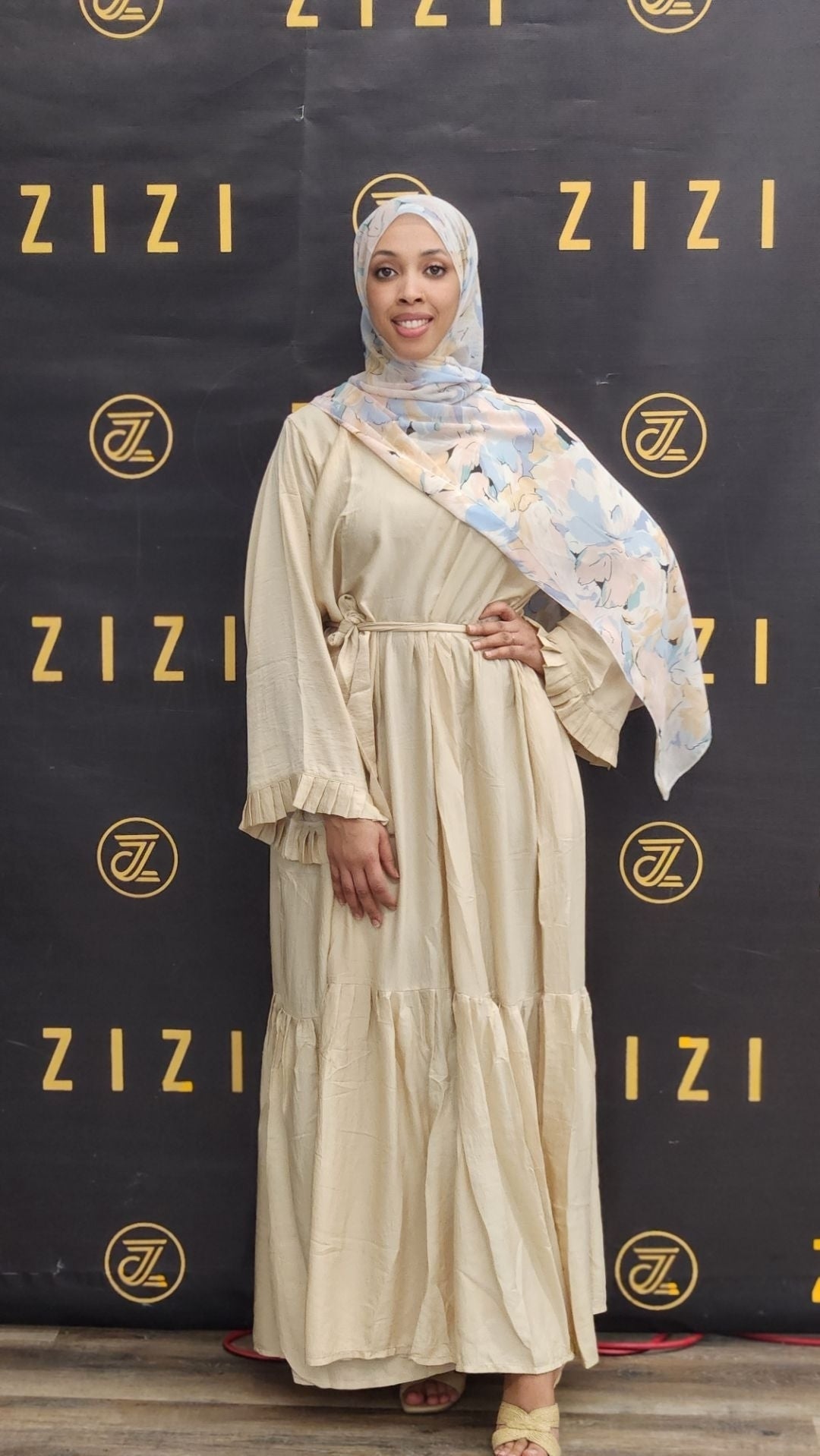Woman in a beige dress with a floral headscarf standing in front of a ZIZI branded backdrop.