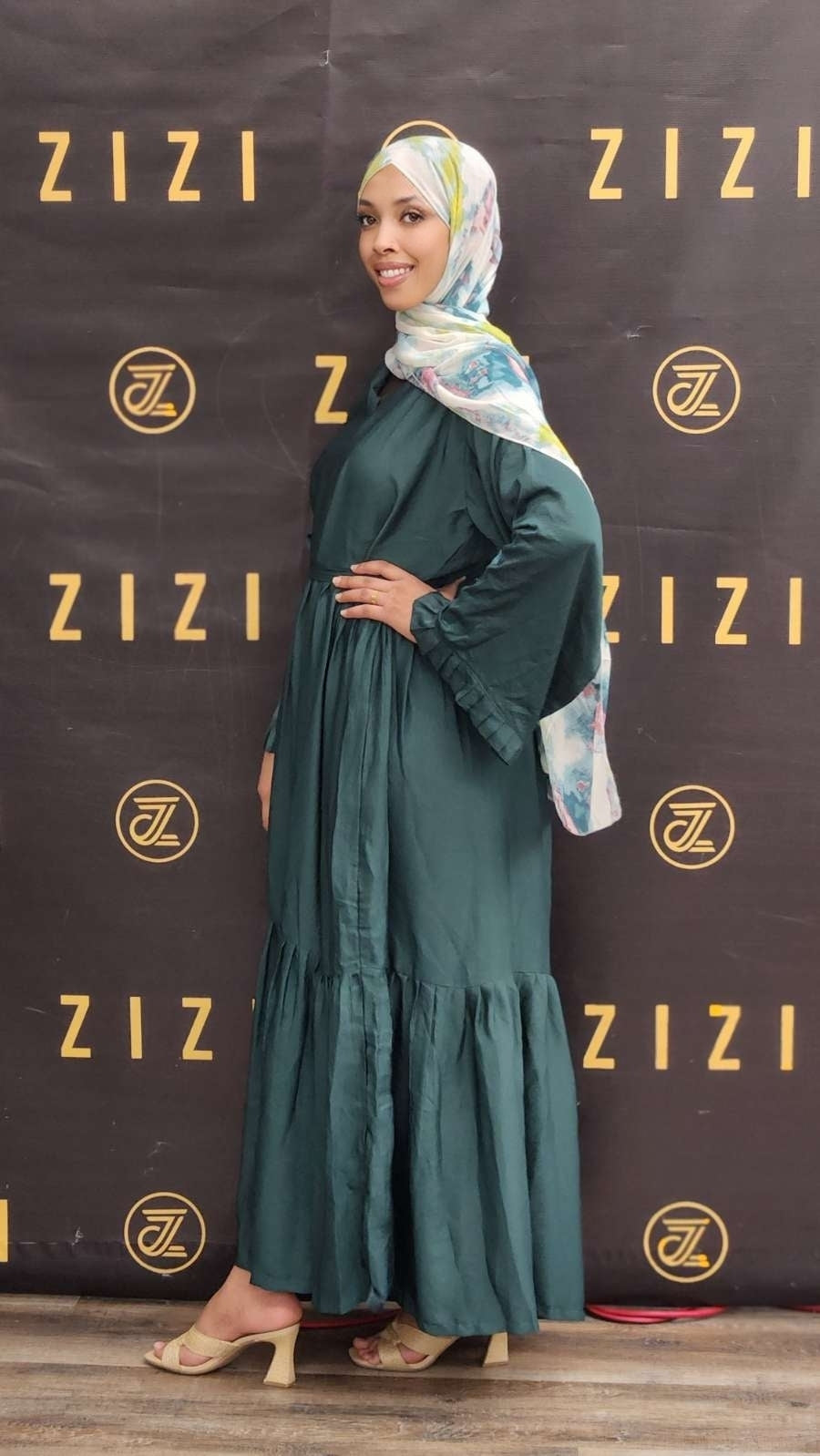 Woman in a green dress and headscarf standing in front of a ZIZI branded backdrop.