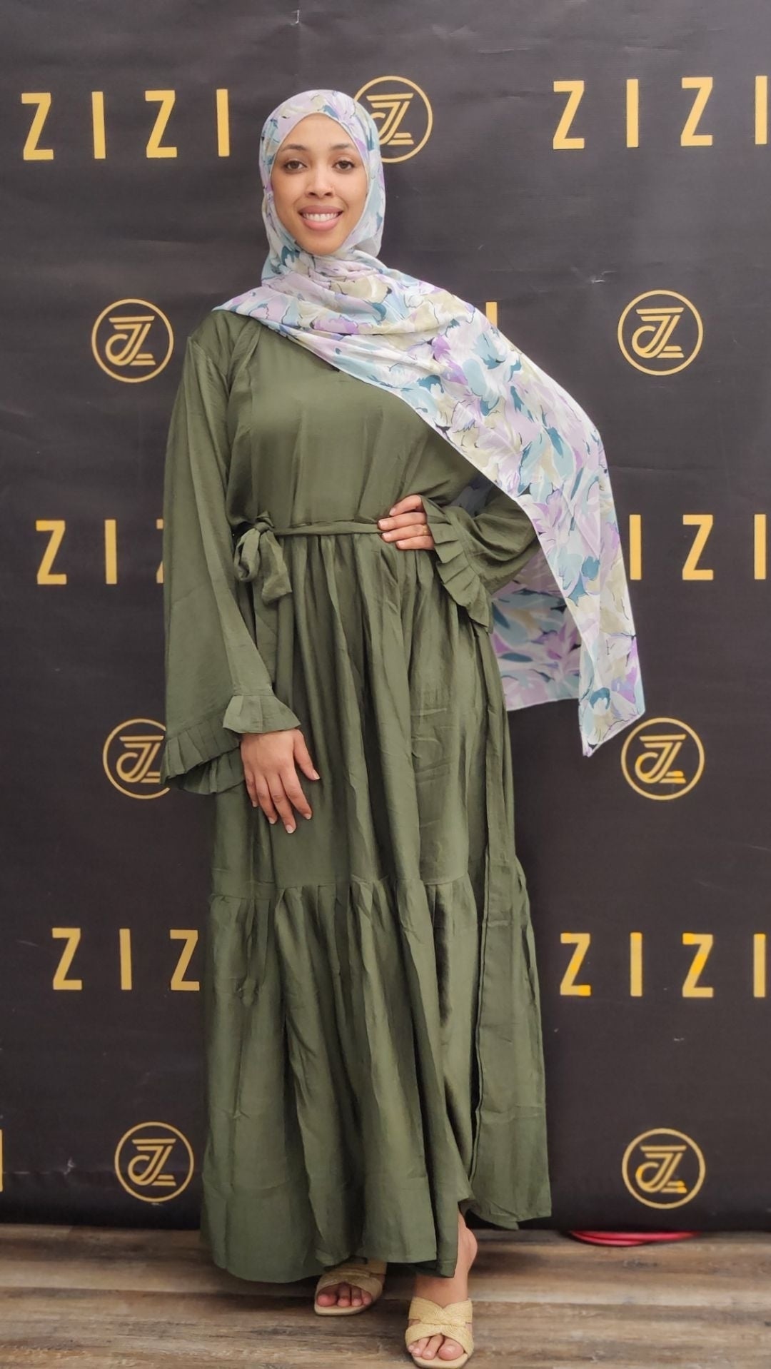 Woman wearing a green dress and floral hijab in front of a Zizi branded backdrop