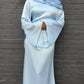 Person wearing a light blue hijab and dress against a dark brick wall.
