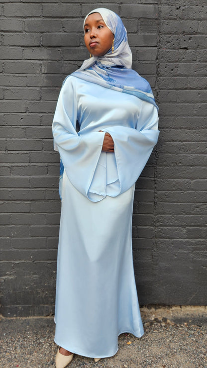 Person wearing a light blue hijab and dress against a dark brick wall.